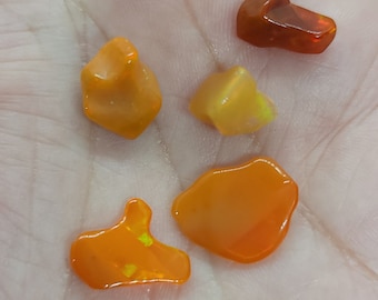 May include: Five polished opal gemstones in shades of orange and yellow are displayed in a hand. The opals have a smooth, glossy surface and varying shapes, with flashes of color visible within the stones. The gemstones are a vibrant display of natural beauty.