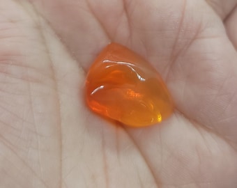 May include: A translucent, orange gemstone with a smooth, polished surface. The gem is teardrop-shaped and appears to be held in a hand. The color transitions from a bright orange to a lighter, almost yellow hue.