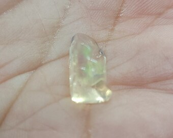 May include: A small, translucent, light yellow gemstone with iridescent flashes of green and yellow. The stone has a rough, irregular shape and appears to be held in a hand. The gemstone is likely a crystal or mineral specimen.