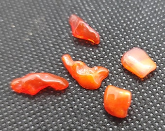 May include: Five polished, translucent, orange-red gemstones of varying shapes and sizes. The stones have a glossy finish and are scattered on a dark, textured surface. The gemstones appear to be natural.