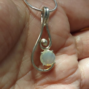 May include: A silver pendant necklace featuring a teardrop design with a spiral accent. The pendant holds a round, iridescent gemstone. The necklace is displayed against a neutral background.