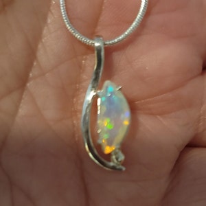 May include: A silver pendant necklace featuring a pear-shaped opal gemstone. The opal displays iridescent colors of green, yellow, and blue. The pendant is suspended from a delicate silver chain, creating an elegant jewelry piece.