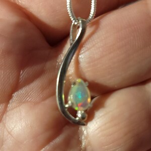 May include: A silver pendant necklace featuring a pear-shaped opal gemstone. The opal displays iridescent colors of green, red, and blue. The pendant is suspended from a delicate silver chain, creating an elegant jewelry piece.