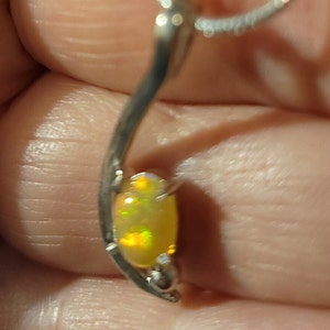 May include: A silver pendant necklace featuring an oval opal gemstone. The opal has a yellow base color with flashes of green and orange. The pendant is suspended from a delicate silver chain.