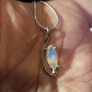 May include: A silver pendant necklace featuring a marquise-cut gemstone with iridescent colors. The pendant is suspended from a delicate silver chain. The gemstone displays a play of blue, yellow, and white hues.