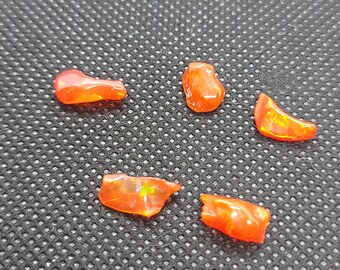 May include: Five polished, orange-red opal gemstones with a fiery, translucent appearance are scattered on a dark, textured surface. The opals vary in shape, showcasing natural formations and internal color play, reflecting light.