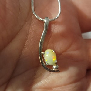 May include: A silver pendant necklace featuring a pear-shaped opal gemstone. The opal has a creamy white base color with flashes of yellow and green. The pendant is suspended from a delicate silver chain.