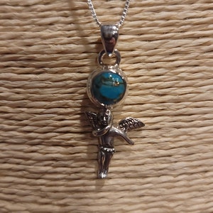 May include: Silver-toned pendant necklace featuring a turquoise-colored stone set in a silver orb, with a detailed cherub figure below. The necklace has a delicate chain and is displayed against a textured, light brown background.