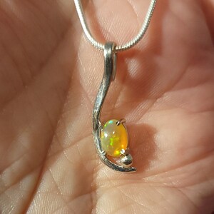 May include: Silver pendant necklace featuring an oval opal gemstone with yellow and green hues. The pendant has a curved, abstract design with a small silver ball accent. The necklace chain is a thin, silver snake chain.