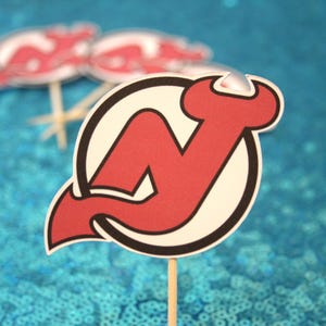 Może przedstawiać: Topper na muffinkę z logo New Jersey Devils w kolorze czerwonym i białym. Logo to stylizowana litera N z czerwonym ogonem diabła.
