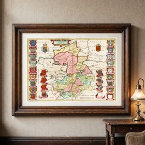 Op de afbeelding: Een ingelijste antieke kaart van Cambridgeshire, Engeland, met de Isle of Ely in roze. De kaart is aan de linker- en rechterkant omzoomd met kleurrijke heraldische wapenschilden, met zwarte tekstlabels die de regionale geografie beschrijven.