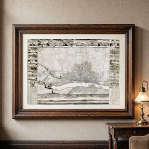 Puede incluir: Un mapa antiguo impreso y enmarcado que muestra un plano cartográfico detallado en blanco y negro de una ciudad junto a un río, rodeado por un borde de pequeñas ilustraciones arquitectónicas, todo dentro de un marco de madera marrón.
