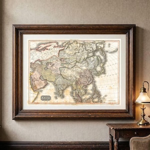 May include: A framed antique-style map of Asia featuring muted earth tones, detailed topographical lines, and geographical labels. The map is enclosed in a dark brown wooden frame with a white mat, displaying the text ASIA in a central box.