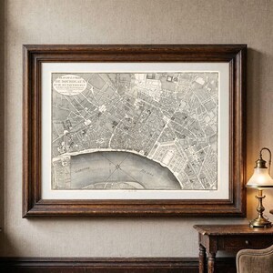 May include: A framed antique map titled Plan de la Ville de Bordeaux et de ses Faux-Bourgs, featuring detailed black and white street layouts and the Garonne River. The map is encased in a dark brown wooden frame with a wide off-white mat border.