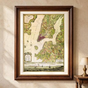 Op de afbeelding: Een ingelijste historische kaart van New York City met de North Hudson River en de East River. De kaart toont gedetailleerde landtopografie in groen- en bruintinten, met een decoratieve cartouche en een panoramisch uitzicht op schepen onderaan.