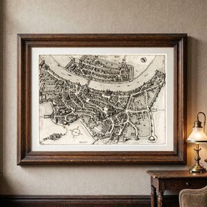 May include: A framed black and white historical map print depicting a city layout with a river, buildings, and fortifications. The artwork is enclosed in a dark brown wooden frame with a white mat border, showcasing intricate line work and details.