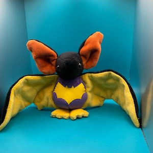 May include: A plush bat toy with yellow wings, a purple body, and a yellow bat symbol on its chest. The bat has orange ears and black eyes.