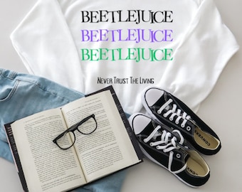 Puede incluir: Sudadera blanca con el texto "Beetlejuice" repetido tres veces en negro, morado y verde. El texto "Never Trust The Living" está impreso debajo en negro. La sudadera está sobre una superficie de mezclilla azul con un libro y zapatillas negras.
