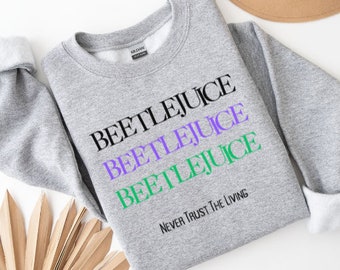 Puede incluir: Sudadera gris con las palabras "Beetlejuice" repetidas tres veces en negro, morado y verde. El texto "Never Trust The Living" está impreso en negro debajo de las palabras repetidas.