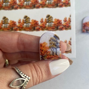 May include: A small, arched-top decorative piece featuring a landscape of autumn trees in shades of orange, yellow, and brown. The piece is held in a hand, with additional landscape strips visible in the background. The design evokes a fall theme.
