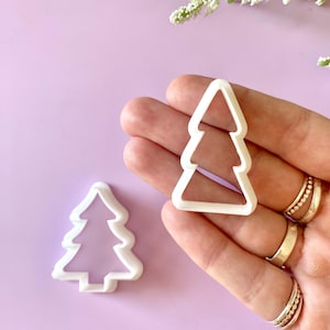 May include: Two white plastic Christmas tree-shaped cookie cutters. One cutter is held in a hand, and the other is on a light purple surface. The cutters have a simple, stylized design, perfect for holiday baking.