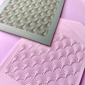 May include: Two square, Art Deco-style molds. One is a light gray with a gold-colored fan pattern. The other is a light purple with a raised, white fan pattern. The molds are likely for crafting or decorative purposes.