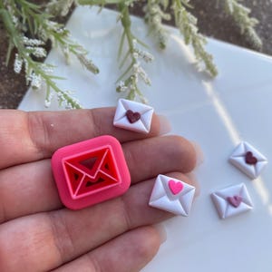 May include: A collection of miniature, heart-themed envelope charms in pink and white. One large pink square envelope charm and three smaller white envelope charms with pink or red hearts. The charms are displayed on a light background.
