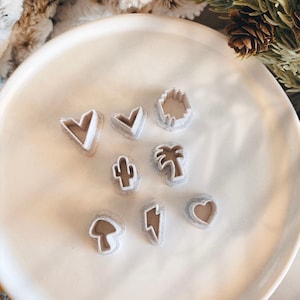 May include: A collection of small, light-colored polymer clay cutters in various shapes, including hearts, a cactus, a palm tree, a mushroom, a lightning bolt, and a sun. The cutters are arranged on a white plate, ideal for crafting projects.