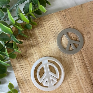 May include: Two peace sign shaped objects. One is white and the other is gray. The peace signs are on a wooden surface with green leafy sprigs in the background. The peace signs are round with the peace symbol in the center.
