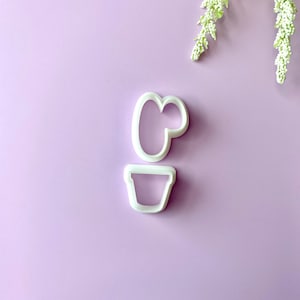 May include: Two white plastic cookie cutters in the shape of a potted cactus, set against a light purple background. The top cutter forms the cactus, and the bottom cutter is a pot. White floral sprigs are in the upper right corner.