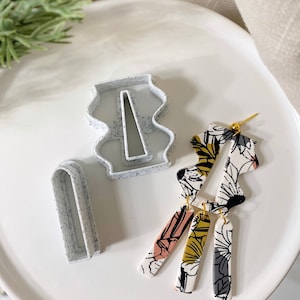 May include: A set of polymer clay earring components and cutters. The earrings feature a floral pattern in black, white, pink, and gold. Two gray cutters with black speckles are also present, along with a white plate.