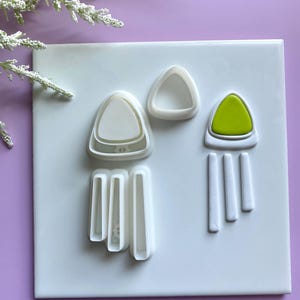May include: A collection of white plastic cookie cutters in various shapes, including triangles and rectangles. One cutter features a green insert. The cutters are arranged on a white surface against a purple background.