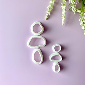 May include: White polymer clay cutters in organic shapes, arranged in two stacks on a lavender surface. The cutters are for crafting and are next to a sprig of white flowers.