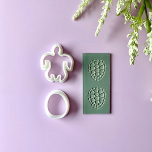 May include: White plastic cookie cutters in the shapes of a person, an oval, and a textured mold with two circular designs. The items are arranged on a light purple surface with white floral accents.