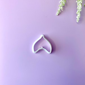 May include: A white plastic petal-shaped cookie cutter. The cutter is on a light purple surface, with a sprig of white flowers in the upper right corner. The cutter is a kitchen tool for baking.