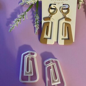 May include: Geometric white and tan earrings with gold hoops, displayed on a white card with the text "LUNARLOVE Design Co." Two matching earring cutters are also shown. The earrings have a modern, abstract design.