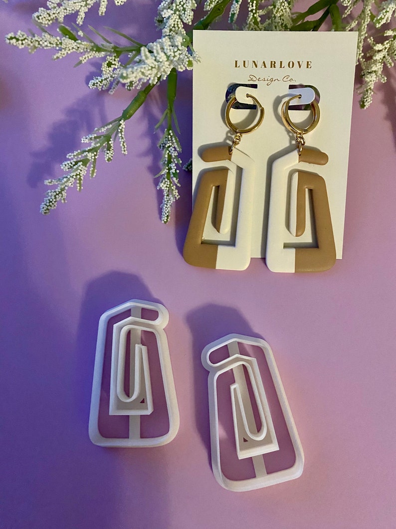 May include: Geometric white and tan earrings with gold hoops, displayed on a white card with the text "LUNARLOVE Design Co." Two matching earring cutters are also shown. The earrings have a modern, abstract design.