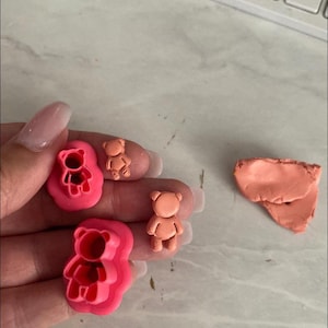 May include: Pink teddy bear clay cutters and small clay bears in various stages of completion. A small piece of orange clay is also visible. The cutters are in the shape of a teddy bear.