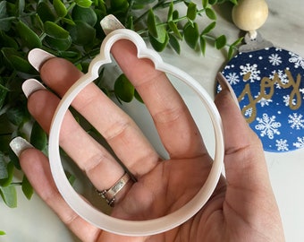 May include: A white plastic ornament-shaped cookie cutter held in a hand. The cutter has a scalloped top and a circular interior. A blue and white ornament with the word "joy" is visible in the background.