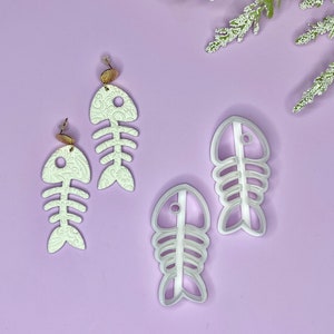 May include: White fish bone-shaped earrings with gold-colored studs and matching polymer clay cutters. The earrings and cutters are on a light purple surface, with white floral accents in the background.