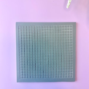 May include: A square, gray silicone mold with a grid pattern. The mold is made of a flexible material and is designed for creating small, square-shaped items. The background is a solid lavender color.