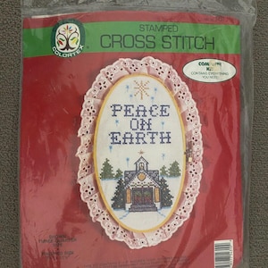 May include: A red and white cross stitch kit with a pink lace trim. The kit includes everything you need to create a cross stitch design that says "Peace on Earth" with a church and trees in the center. The kit is packaged in a clear plastic bag.