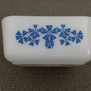 May include: A rectangular, white Pyrex dish with a blue floral pattern. The dish has rounded corners and a slightly flared rim. The blue design is symmetrical and features stylized flower-like shapes. The dish is likely vintage and suitable for kitchen use.