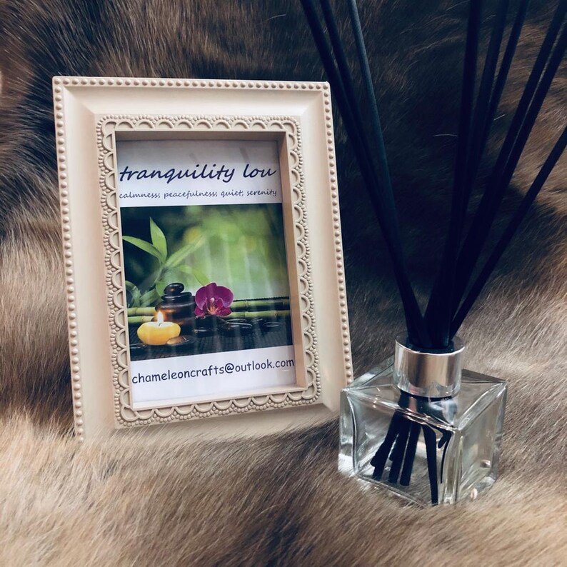 May include: A white picture frame with a nature scene and the text "tranquility low calmness, peacefulness, quiet, serenity" and the email address "chameleoncrafts@outlook.com". A clear glass bottle with black diffuser sticks is in the foreground.