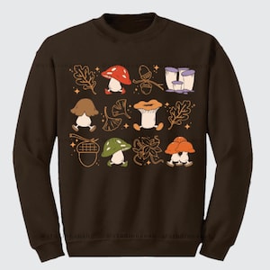 Peut inclure: Sweat-shirt marron avec un motif coloré de champignons et de feuilles. Les champignons sont de formes et de tailles variées, et les feuilles sont dans des tons de vert et de brun.