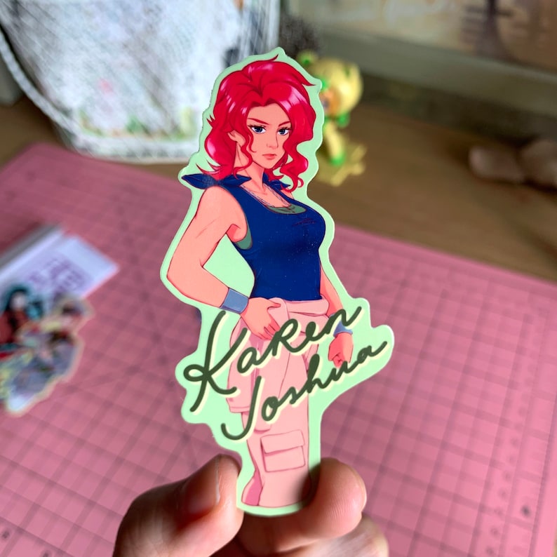 May include: A sticker featuring a cartoon woman with red hair wearing a blue tank top and khaki pants. The sticker has a green background and the text "Karen Joshua" is written in cursive.