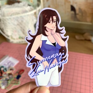 May include: A sticker featuring a cartoon illustration of a woman with long brown hair wearing a blue and white top and white pants. The sticker has a purple background and the text "Summeragi Lee Nariega" in blue.