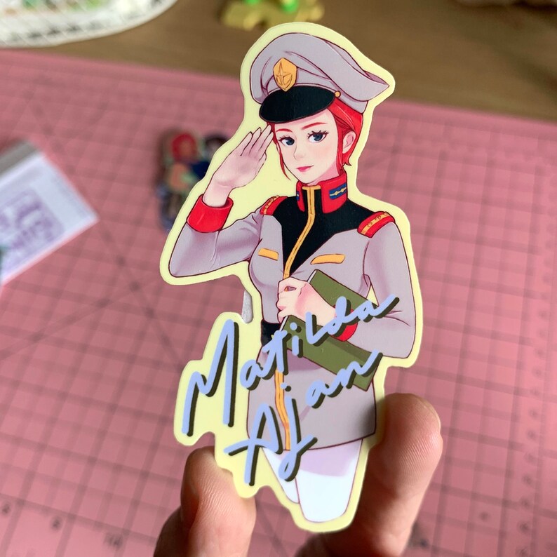 May include: A sticker featuring a cartoon woman in a military uniform with a red and white hat, saluting. The sticker has a yellow background and the text "Matila Ajian" in blue and yellow lettering.