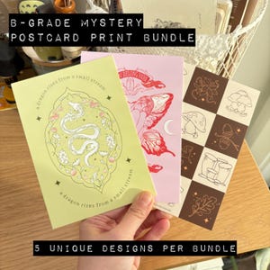 May include: A selection of B-Grade mystery postcards with unique designs. The postcards showcase illustrations of a dragon, a butterfly, and other nature-inspired images. Text on the postcards includes "B-GRADE MYSTERY POSTCARD PRINT BUNDLE", "a dragon rises from a small stream", and "5 UNIQUE DESIGNS PER BUNDLE".