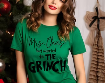 Puede incluir: Una camiseta verde con el texto "Mrs. Claus but married to THE GRINCH" en negro. La camiseta es de cuello redondo. El fondo incluye un árbol de Navidad y decoraciones. La persona lleva la camiseta.